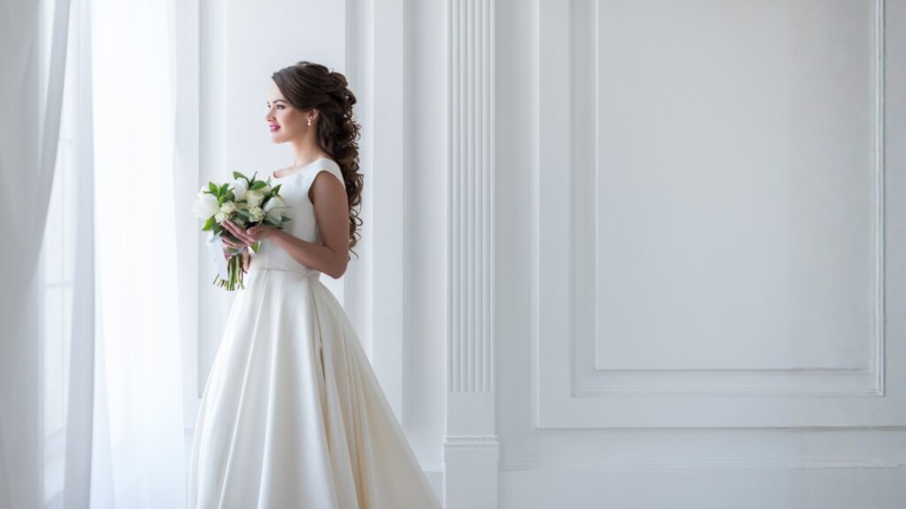 Novia elegante de perfil junto a una ventana brillante sosteniendo un ramo, imagen de portada sobre tratamientos faciales para bodas en Murcia, Clínica Lorents